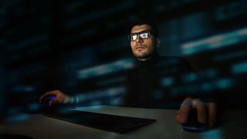 A Man Using a Computer in A Dark Room with Data Reflected on His Glasses, Representing the Concept of Anonymity Online