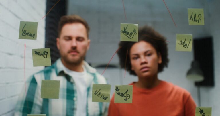 A man and a woman stand behind a glass wall covered in sticky notes, intensely brainstorming and planning for a startup in Finland