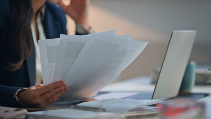 A business translator in a professional suit reviewing multiple printed documents at a desk, with a laptop open in front of them