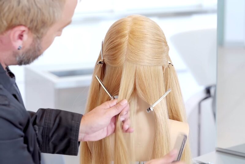 A hairstylist demonstrating a technique on a mannequin head with long blonde hair, sectioned with clips, in a bright salon