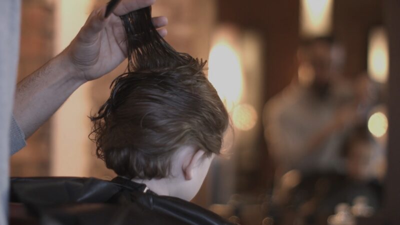 A hairstylist lifting and trimming a young child's damp hair inside a salon, with a blurred background of other clients