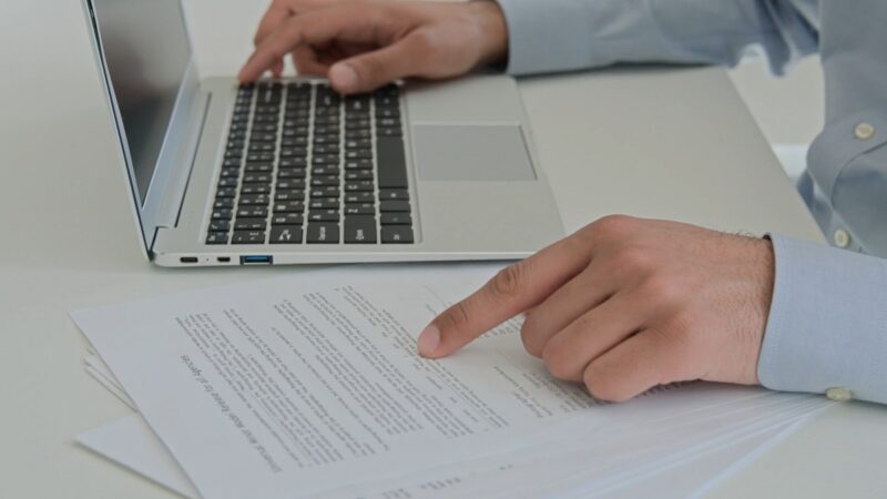 A man in a dress shirt pointing at a printed document while working on a laptop