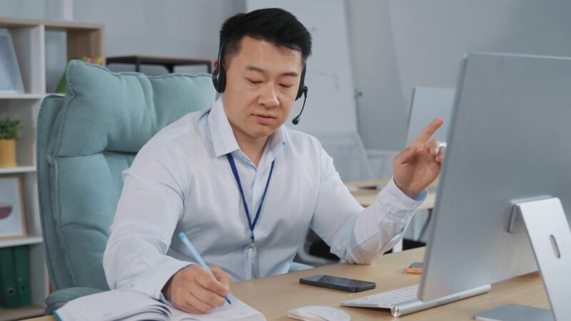 A professional man wearing a headset and business attire, taking notes while speaking in an online meeting